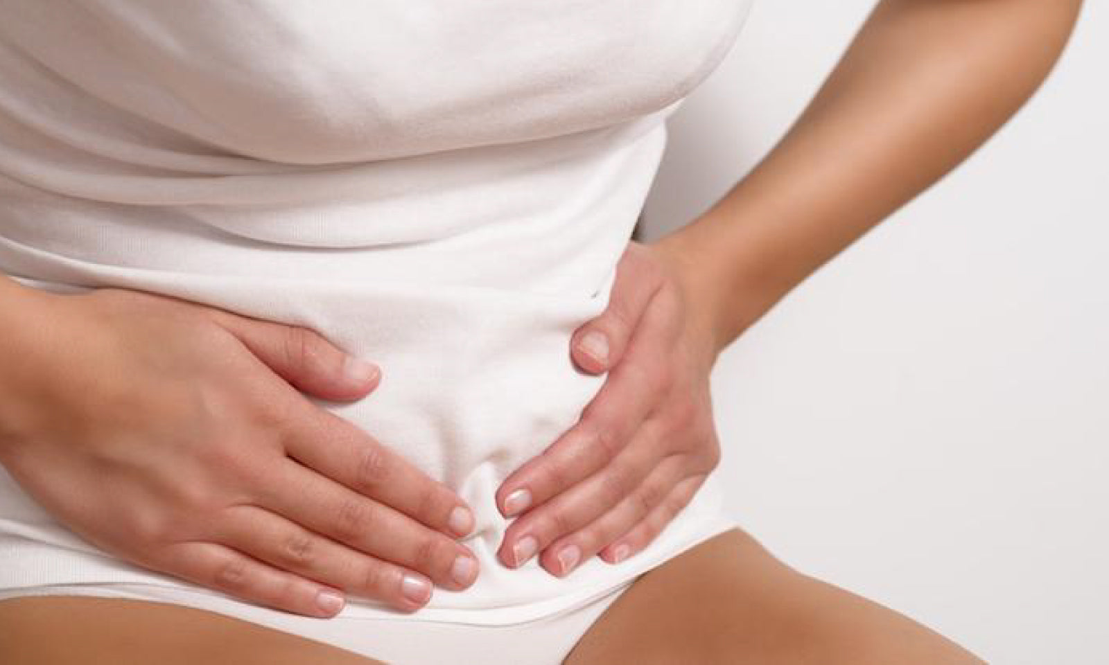 A woman in a white t-shirt and white underwear presses both hands to her stomach.