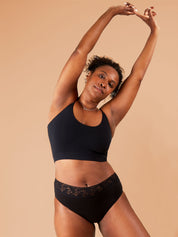 A brown-skinned woman standing and looking directly at the camera with a content look, raising her hands in the air as if stretching. She is wearing black period pants with floral lace trim and a cropped bra.