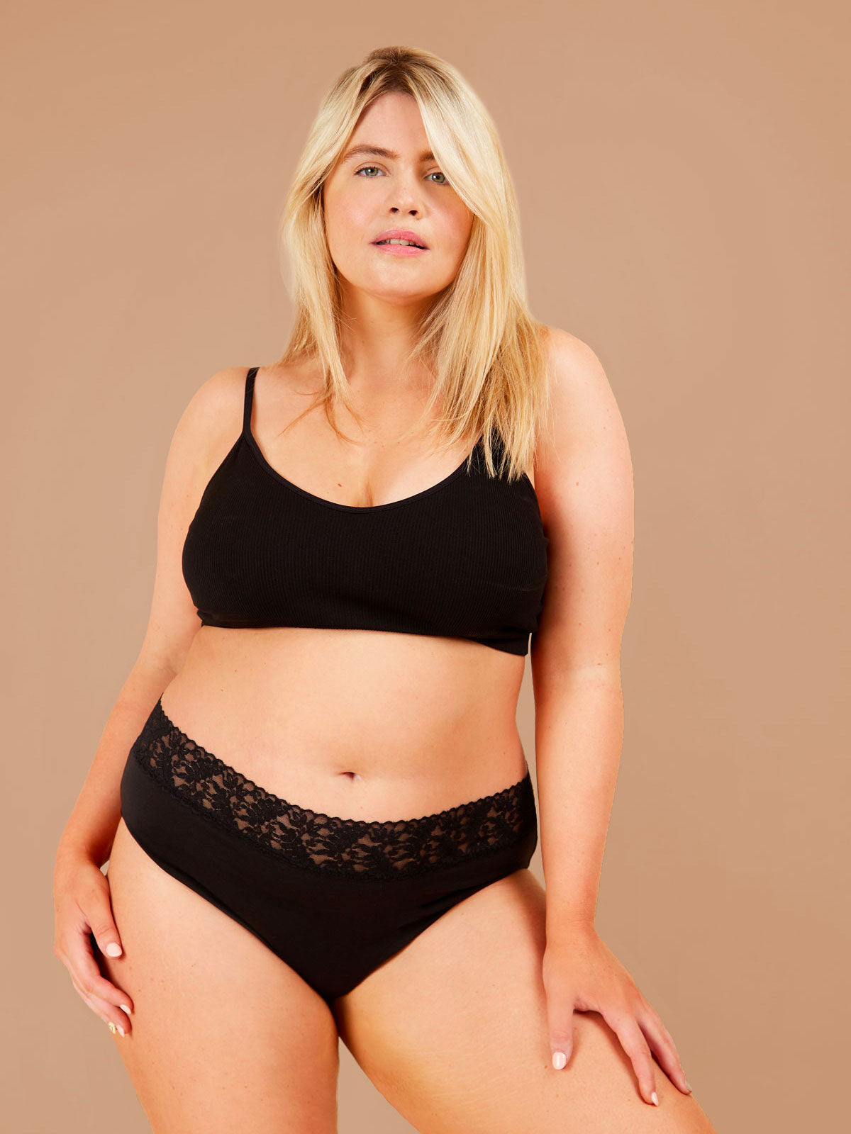 A blonde woman stands wearing a black bra and black period pants adorned with a lacey floral trim, looking content and directly at the camera.